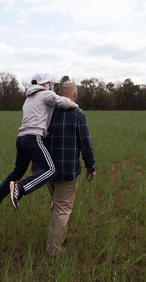 A man carrying a boy child piggy-back style in a field of green.