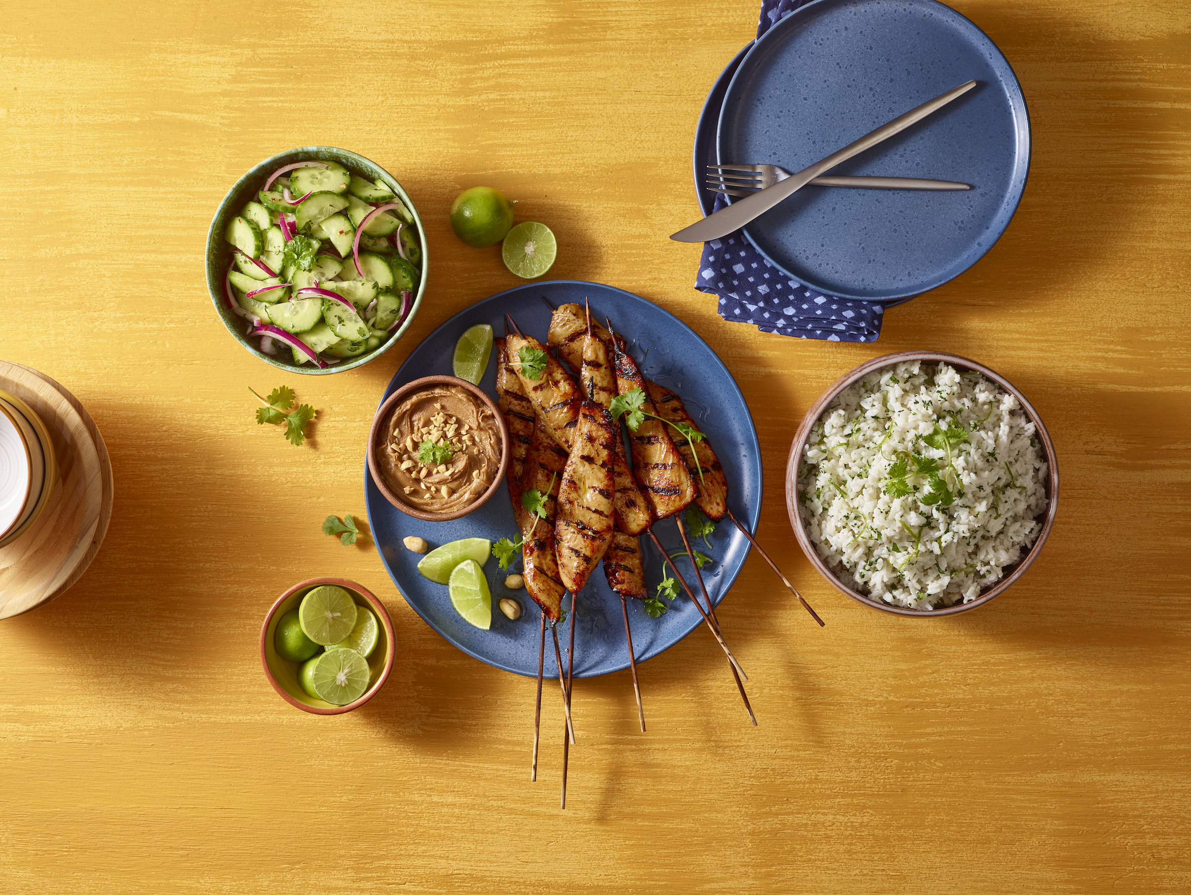A meal with grilled turkey satays, rice, cucumber salad, peanut sauce and lime on a wooden table.