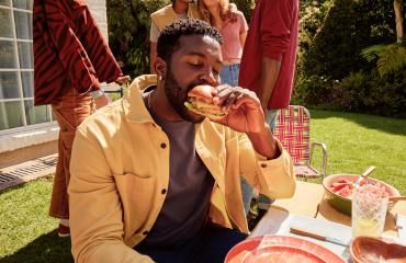 Man eating a turkey burger at an outdoor gathering with friends on a sunny day