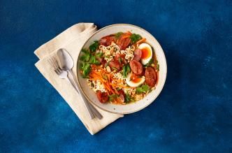 Bowl of ramen with spicy turkey sausage, eggs, carrots, cilantro, and peanuts on a blue background.