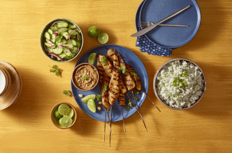 A meal with grilled turkey satays, rice, cucumber salad, peanut sauce and lime on a wooden table.