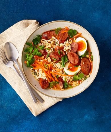 Bowl of ramen with spicy turkey sausage, eggs, carrots, cilantro, and peanuts on a blue background.